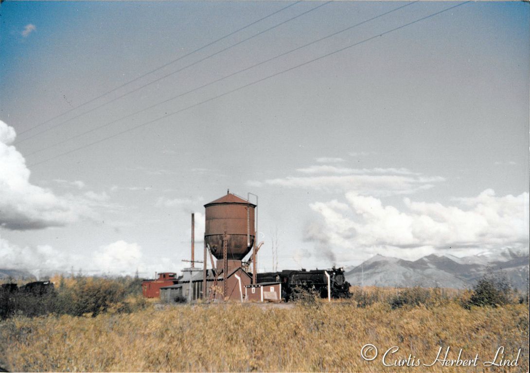 Matanuska water tank, the only one with two spouts. The 550 class locomotive is on the Palmer/Chicaloon branch and the other 550 class locomotive to the left is on the same track. The main line to Wasilla and up to Fairbanks is in the foreground of the tank. Photo dates after 1944 and before 1958. Fall colors indicate September or October and the view is North East with Lazy Mountain in the background. 