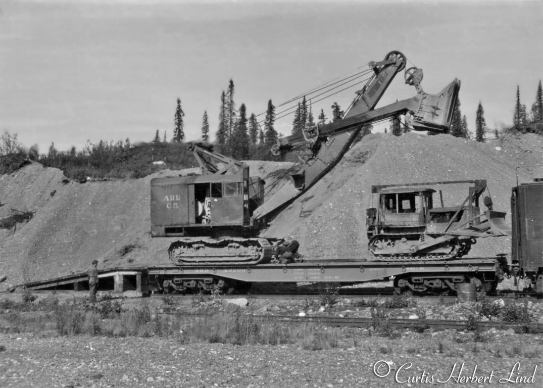 Here they are preparing the ARR C5 shovel for a short move. We can hope it is a short move if they plan to leave the boom and bucket up in the air. This machine and the cat are both cable operated, no hydraulics. The flat at least is a heavy duty car with 6 wheel trucks, but with both vehicles setting over the trucks it still a marginal load. Notice there are two people on the car deck probably setting chocks. The cut bank in the back ground appears to be the source of the gravel being loaded out.Great shot of X-3200 75 ton capacity heavy duty flatcar Built for the US Army Transportation Corps. Acquired in 1950 by the ARR as 2nd #5570, became work car X-3200.